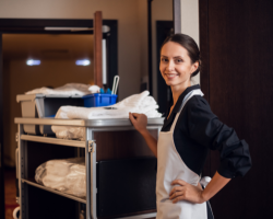 A woman in a smock pushes a cleaning trolley and smiles at the camera