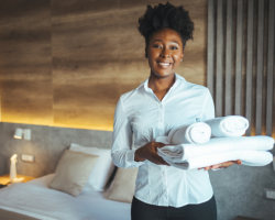 Woman with a white blouse carries towels in her arms and stands in a tidy hotel room