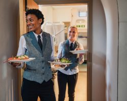 Two waiters in vests and ties carry plates of food from the kitchen