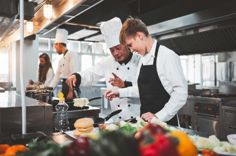An older chef shows a younger chef how to prepare a dish in a large kitchen