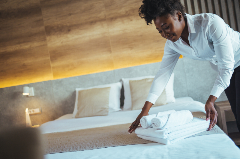 A woman in a white blouse lays towels on a made-up bed in a hotel room
