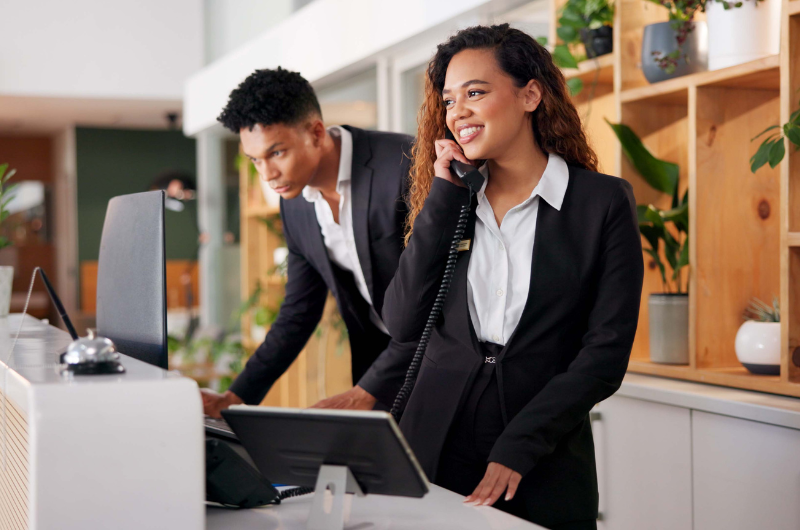 Eine Frau und ein Mann stehen an einer Rezeption, die Frau telefoniert und der Mann schaut auf den Computer