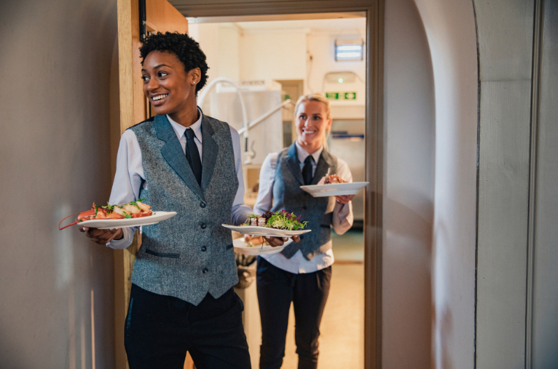 Two waiters in vests and ties carry plates of food from the kitchen
