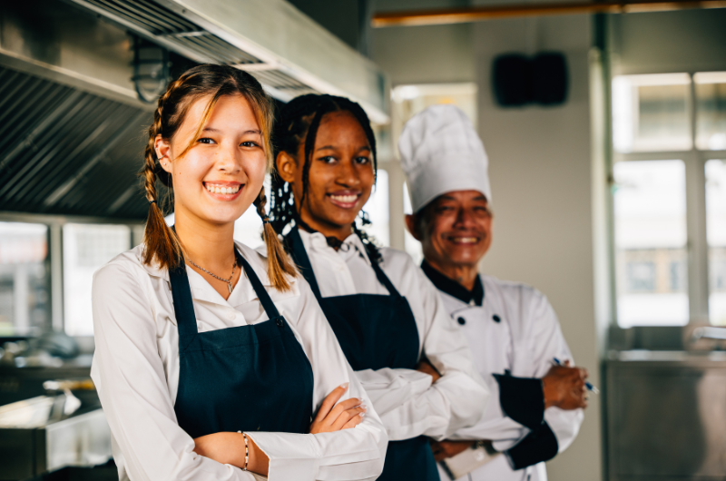 Two female and one male kitchen staff in aprons smile into the camera